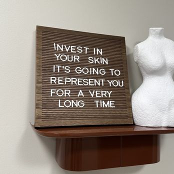 Defined - A rectangular wooden sign with horizontal grooves features a quote in white capital letters: 'INVEST IN YOUR SKIN, IT'S GOING TO REPRESENT YOU FOR A VERY LONG TIME.' The sign is placed on a brown shelf attached to a plain, light-colored wall. To the right of the sign, there is a white mannequin torso without arms or head, made of textured material, also resting on the same shelf. - Medical Spas Near Me - Chicago, Illinois
