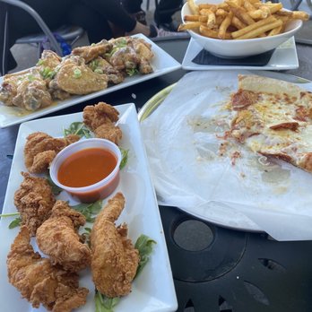 Flashback Retro Arcade + Bar + Grille - Chicken Tenders with a side of buffalo, Michelangelo pizza with pepperoni, Cajun fries, Garlic Parm wings - The image shows a table with multiple dishes including fried chicken wings with a side of dipping sauce, a partially eaten slice of pizza, a bowl of French fries, and a plate of chicken with sauce. The setting appears to be an outdoor dining area with chairs and people seated in the background. The food presentation suggests casual dining, possibly at a restaurant or bar with outdoor seating. - Bars Near Me - Hyannis, Massachusetts