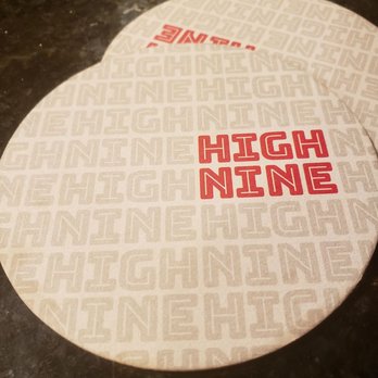 High Nine Brewing - Two round paper coasters with the words "HIGH NINE" printed in red are positioned on a dark, reflective table surface. The coasters have a repeating pattern of the same text in a lighter color as background. The setting appears to be a bar or brewery environment. - Breweries Near Me - Deep River, Connecticut