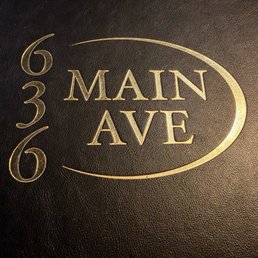 A photograph features a black textured menu cover with gold embossed lettering. The text includes the address '636 Main Ave' arranged vertically on the left side and 'Main Ave' inside an oval shape on the right. The design is simple and elegant, indicating it is likely from a restaurant or similar establishment located at that address.