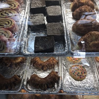 La Cosecha - a variety of baked goods - A glass display case showcases an assortment of baked goods and confections. On the left, there are rolled pastries with a spiral pattern, possibly cinnamon rolls or similar. In the center, there are several dark chocolate or fudge squares arranged in rows. To the right, there are croissants or similar pastries with a golden-brown crust. At the bottom, there are crescent-shaped pastries and a cookie decorated with colorful sprinkles. The items are placed on aluminum foil, and the display is behind a glass surface, with reflections visible. - Empanadas Near Me - Somerville, Massachusetts