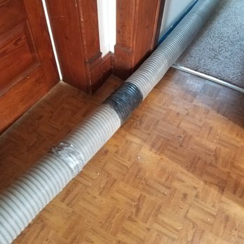 Critter Control - More dust leaking out. - A flexible, ribbed duct with black soot marks is placed on a wooden floor near a wooden door and a carpeted area. - Pest Control Near Me