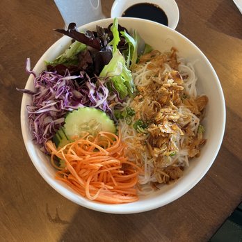 Bamboo by Kulantro - A bowl of Vietnamese noodle salad contains shredded purple cabbage, sliced cucumber, shredded carrots, and mixed greens, along with rice noodles topped with grilled meat and fried shallots. A small dish of soy sauce is placed nearby on a wooden table, with a tall glass and utensils partially visible. The presentation emphasizes fresh ingredients and diverse textures. - Vietnamese Near Me - Albuquerque, New Mexico