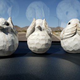 Three white rabbit figurines, carved with textured details, are arranged on a car dashboard. The background shows a desert landscape with mountains and a clear blue sky, visible through the windshield. The figurines are evenly spaced and face forward, with reflections of the sky and clouds visible on the windshield.