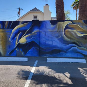 Lively Mind Art Studio - A mural on a concrete wall depicts a hand holding a paintbrush, with smoke or cloud-like shapes in the background, in an outdoor parking area. - Arts & Crafts Near Me - Phoenix, Arizona