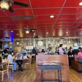 Mo's Egg House - The image shows the interior of a restaurant with a red ceiling featuring ceiling fans and recessed lighting. Diners are seated at wooden tables and booths, some wearing masks, engaged in eating or conversation. The back wall has a large clock, wall art, and decor spelling 'MOO'. The floor is wooden, and staff are visible serving or attending to customers. The overall setting appears casual and family-friendly. - Breakfast & Brunch Near Me - Temecula, California