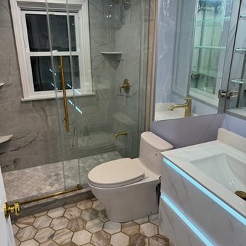 Sun Shore Construction - A bathroom features a glass shower with brass hardware, a white toilet, and a vanity with a sink and illuminated mirror. The floor has hexagonal tiles. - General Contractors Near Me - Needham, Massachusetts