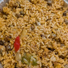Arroz Con Gandules - The image shows a round, metallic container filled with cooked rice mixed with vegetables such as red and green peppers and beans. The rice appears seasoned and cooked thoroughly, with visible pieces of vegetables distributed throughout. The container is partially visible, and the dish looks like a prepared rice meal, possibly for takeout or serving at a gathering.