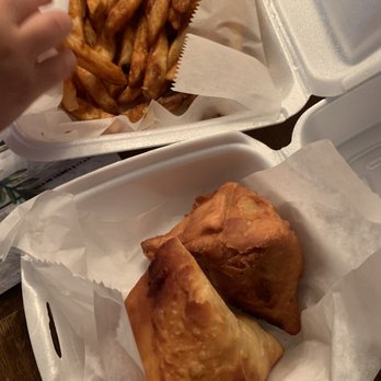 Yumy Platter - French fries & samosas - The image shows two foam takeout containers with food from a Mediterranean-inspired eatery. One container holds a portion of French fries, and the other contains two samosas on white paper. The fries are golden-brown and crispy, while the samosas are deep-fried with a golden exterior. The containers are placed on a dark surface, and a hand is reaching into the fries container. The setting suggests a casual takeout meal. - Seafood Near Me - Wrightstown, New Jersey