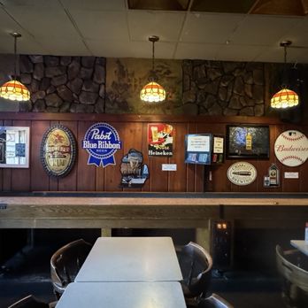 Stober's - The image shows the interior of a bar with wood-paneled walls decorated with various beer signs, including Budweiser, Pabst Blue Ribbon, and Heineken. A woman is seen setting up or cleaning a shuffleboard table on the left side. The bar has a dark, cozy atmosphere with hanging pendant lights and a stone accent wall above the paneling. Tables and chairs are arranged in the foreground, and the overall setting suggests a casual, community-oriented bar environment. - Bars Near Me - Lansing, Michigan