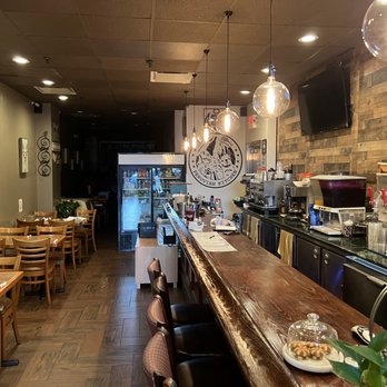 Amar Peruvian Kitchen - Indoors - The photo depicts the interior of a Peruvian restaurant featuring a long wooden bar counter with bar stools. To the left, there are several wooden tables with chairs arranged for dining. The back wall has a logo reading 'Peruvian Kitchen' and a refrigerator visible through an open doorway. The right side shows kitchen equipment, including a coffee machine and beverage dispensers, with a wood-paneled wall and mounted TV screens. The ceiling has hanging light fixtures, and the overall decor combines wood accents and neutral tones. - Peruvian Near Me - Forest Hills, New York