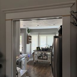 Door trim - The image shows a doorway leading into a kitchen with white plantation shutters, wooden flooring, and various kitchen items on a cart and counter.