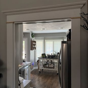 The Unhinged Door - Door trim - The image shows a doorway leading into a kitchen with white plantation shutters, wooden flooring, and various kitchen items on a cart and counter. - Door Sales/Installation Near Me