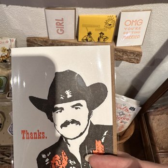 Luna and Luz - Burt! (6/13/2023) - A greeting card displays a black-and-white stylized portrait of a man wearing a cowboy hat and mustache, with the word "Thanks." in red. It is held by a person in a store with shelves of other cards and items. - Jewelry Near Me - Albuquerque, New Mexico