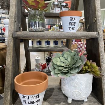 Mavens & Makers - a shelf with potted plants - A wooden shelf holds two decorative plant pots with succulents, one with a message reading 'TALK DIRTY TO ME' and the other saying 'YOU MAKE ME WANNA SPROUT.' The pots are terracotta and white ceramic, respectively. Behind them, there are small jars and candles, with a blurred background of a store interior. The arrangement emphasizes rustic and playful decor elements. - Gift Shops Near Me - Cullman, Alabama