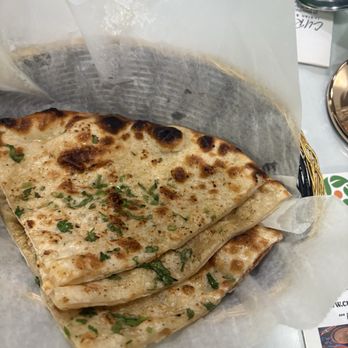 Curry Leaf - Garlic naan - A close-up of garlic naan bread with chopped herbs on top, resting on parchment paper inside a basket. The naan has a golden-brown, slightly charred surface, indicating it was baked in a tandoor or oven. The background shows a table setting with a spoon, a menu, and a small dish, suggesting a restaurant environment. - Indian Near Me - Foxborough, Massachusetts