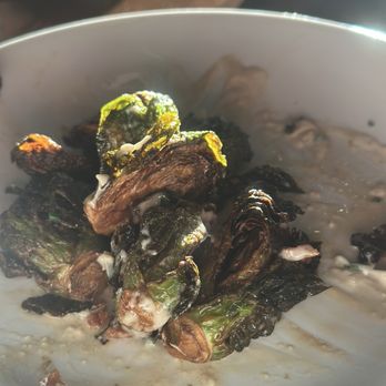 Chomp Kitchen and Drinks - Salt and vinegar Brussels - A white bowl contains charred Brussels sprouts with a dark, crispy exterior, topped with a creamy sauce. The bowl is placed on a wooden surface, with a blurred background showing a smartphone and other objects. The image emphasizes the roasted texture of the Brussels sprouts and the creamy topping. - Burgers Near Me - Newport, Rhode Island