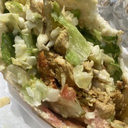 Chicken Shawarma - A close-up image of a chicken shawarma pita sandwich showing shredded lettuce, chopped vegetables, and grilled chicken pieces inside a pita bread. The pita is partially open, revealing the filling, and the background includes aluminum foil. The sandwich appears freshly prepared, with a variety of textures and colors from the vegetables and meat.