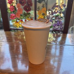 Black Bear  Latte (Orange Mocha) - A latte in a paper cup with a slice of dried orange on top is placed on a wooden table near a window with colorful floral stained glass.