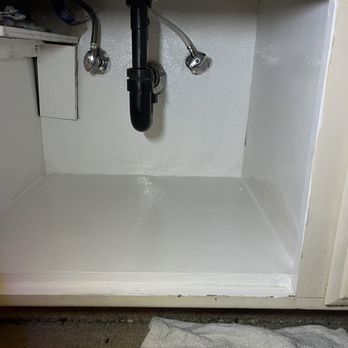 Same Day Handyman - After - The photo depicts the interior of a cabinet beneath a kitchen sink, featuring a black drain pipe and flexible water supply lines with metal fittings. The cabinet walls and floor are painted white, and the area appears clean and freshly painted. Part of a countertop and a towel on the floor are also visible. - Handyman Near Me