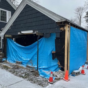 Verndex - The image shows a house with part of its exterior wall being renovated. Blue tarps are draped over the side of the house, covering the area where siding has been removed, exposing the underlying wooden framing. Construction cones are placed nearby, and the surrounding area has some snow on the ground. The house has dark-colored siding and a gabled roof, with neighboring houses and leafless trees visible in the background. - General Contractors Near Me - Madison, Ohio