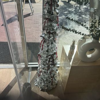 JULIA - A tall, silver artificial Christmas tree decorated with red and black ribbons, ornaments, and other embellishments is situated indoors near a glass door. The tree is placed on a wooden floor next to a light-colored wooden table with decorative items, including a white vase with greenery and a small cone-shaped object. Outside the glass door, benches and a person sitting on a bench are visible, with sunlight casting shadows on the pavement. - Jewelry Near Me - Fresno, California