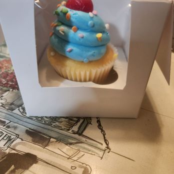 Lulu's Bakery - Vanilla cupcake - A cupcake with blue frosting decorated with colorful sprinkles and a cherry on top, displayed in a white bakery box with a clear window. - Bakeries Near Me - Fresh Meadows, New York