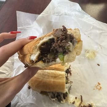 Algonquin Sub Shop - Cheesy Italian Beef with Sweet Peppers - A sandwich with beef, green peppers, and melted cheese is held by a hand with red nails, placed on a paper-lined surface. - Sandwiches Near Me - Algonquin, Illinois
