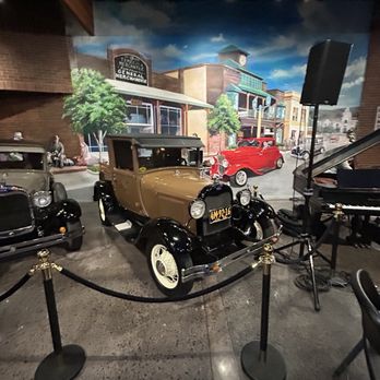 Jack’s Garage - The image shows an indoor exhibit with a vintage automobile, painted beige with black accents and white-walled tires, positioned on a concrete floor. The car is enclosed by a black rope barrier with gold stanchions. Behind the car, there is a mural depicting a street scene with buildings, trees, and classic cars, creating a historical ambiance. To the right, a speaker and a piano are visible, indicating a setting that may include live music or presentations. - Museums Near Me - Temecula, California