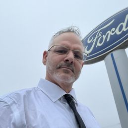 Exterior outside entrance of Newport Ford - Shawn Helmick with ford sign in back - Newport, Oregon Exterior outside entrance of Newport Ford - Shawn Helmick with ford sign in back - Newport, Oregon
