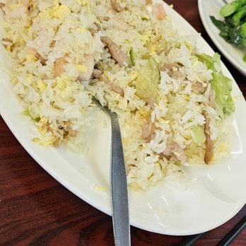 Dumpling House - B17. House Special Fried Rice - A large oval white plate contains house special fried rice with shrimp, pork, and vegetables, including lettuce and egg. A metal spoon rests on the plate. The table has chopsticks and additional dishes, including rice and greens. A red drink and part of a person's arm are visible in the background, indicating a restaurant setting. - Chinese Near Me - Newton, Massachusetts