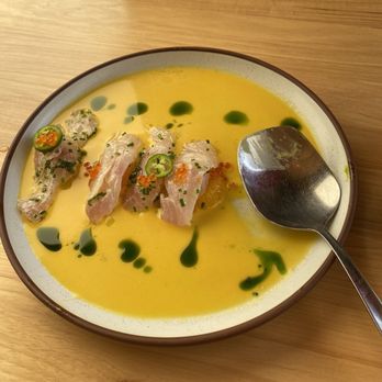 Bulevar Mexican Kitchen - A bowl of yellow-colored soup or sauce with slices of raw fish, jalapeño slices, and green sauce drops on top, placed on a wooden table. - Mexican Near Me - Austin, Texas