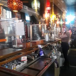 The image shows the interior of a bar with multiple hanging pendant lights of different designs and colors. A bartender is seen on the right side, preparing drinks behind the bar counter. The bar has various equipment, including taps, bottles, and cleaning supplies. A partially visible beer can is in the foreground, and the background includes patrons and additional lighting, creating a lively atmosphere.