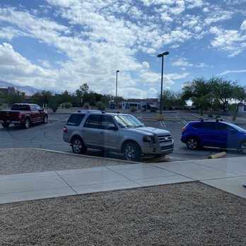 US Post Office - A parking lot scene featuring several parked cars, including a silver SUV in the foreground, a red truck to the left, and a blue car to the right. The lot has designated parking spaces with concrete wheel stops, and there are light poles and some trees in the background. The sky above is partly cloudy with scattered white clouds and patches of blue, suggesting a clear day. - Post Offices Near Me - Tucson, Arizona