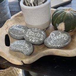 Gray decorative stones with handwritten herb names such as parsley, cilantro, chives, and sage are placed on a wooden tray. A ceramic container filled with metal utensils labeled with herb names is nearby. A pumpkin is also present on the tray, which is set on a wooden surface, with additional metal buckets and a tray visible in the background.