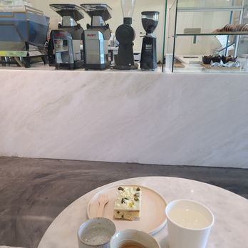 Onescape Coffee - Espresso, tonic water, pistachio chocolate cake, and ice water. The background is the barista's crafting area/counter. - Coffee & Tea Near Me - Los Angeles, California