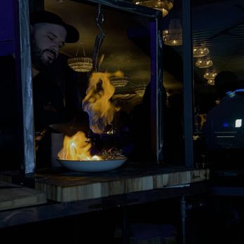 Astra Rooftop - Tomahawk Steak - A chef is preparing a dish with visible flames rising from a bowl, using a cooking setup at Astra Rooftop in Columbus, Ohio. The background shows a dimly lit interior with hanging light fixtures and a reflective ceiling, indicating a restaurant setting. - American Near Me - Columbus, Ohio