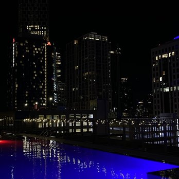 Edge Rooftop - hotel pool right next to Edge - The image shows a city skyline at night with multiple high-rise buildings illuminated by interior lights. A rooftop pool with blue lighting is visible in the foreground, reflecting the city lights. String lights are strung along a railing, adding to the scene's ambiance. The sky is dark, emphasizing the bright lights of the buildings and pool. - Lounges Near Me - Austin, Texas