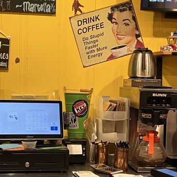 Koco's Pub - A coffee station features a Bunn coffee machine, a large KEG cup, and various supplies including pens, a calculator, and papers. A sign with retro artwork and the text "Drink Coffee" is mounted on a yellow wall, alongside a small figurine and a television screen. The setup appears to be in a casual cafe or diner environment. - Pubs Near Me - Baltimore, Maryland