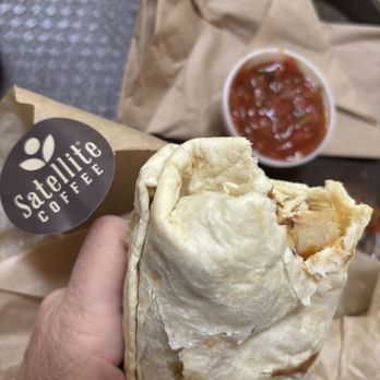 Satellite Coffee - Burrito and salsa - A hand holds a partially eaten burrito wrapped in paper with Satellite Coffee branding, with salsa in a cup and a paper bag in the background. - Coffee & Tea Near Me - Albuquerque, New Mexico