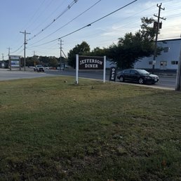 Advertising open when closed - The image shows a sign for Jefferson Diner situated on a grassy area adjacent to a road. The sign is black with white lettering and indicates that the diner is open. Several cars are visible on the road, and there are utility poles and power lines overhead. In the background, there are trees and a building, with a partly cloudy sky above.