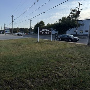 Jefferson Boulevard Diner - Advertising open when closed - The image shows a sign for Jefferson Diner situated on a grassy area adjacent to a road. The sign is black with white lettering and indicates that the diner is open. Several cars are visible on the road, and there are utility poles and power lines overhead. In the background, there are trees and a building, with a partly cloudy sky above. - Breakfast & Brunch Near Me - Warwick, Rhode Island