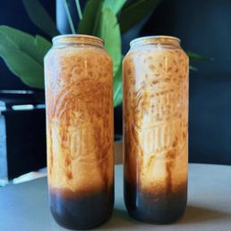 Banana Bread - Two tall cans of iced coffee with a layered appearance, featuring a dark base and caramel-colored top, placed on a table with a plant in the background.