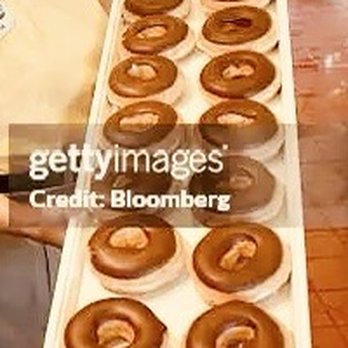 Krispy Kreme - A rectangular white tray displays fifteen glazed donuts, arranged in two rows, with some donuts placed on paper doilies. The donuts are evenly spaced and appear freshly glazed. The background shows a wooden surface and part of a person's leg, indicating an indoor setting, possibly a bakery or donut shop. - Donuts Near Me - Manassas, Virginia