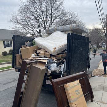 Junk Pro - A truck is parked on a residential street, loaded with various items including furniture, cardboard boxes, and packaging materials. The truck's bed is overflowing with items, some of which are wrapped or boxed, and a large cardboard box labeled 'Fragile Glass' is placed on the sidewalk. The background shows houses, leafless trees, and an overcast sky, indicating a neighborhood setting. - Junk Removal & Hauling Near Me - Newton, Massachusetts
