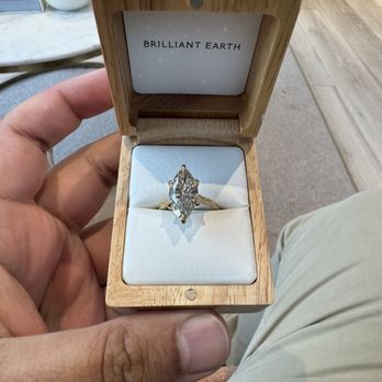 Brilliant Earth - A person holds an open wooden jewelry box displaying a diamond ring with a marquise cut, set in a metal band. The box has a white interior and a label reading 'Brilliant Earth' on the inside of the lid. The background includes a table with a cup, a book, and other furniture, indicating a retail or showroom environment. - Jewelry Near Me - Chicago, Illinois