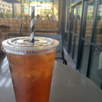 Hawt Pizza Co. - Mint iced tea - A transparent plastic cup filled with iced tea and ice cubes, covered with a clear plastic lid and a black-and-white striped straw, is positioned on a dark table. The background features large glass windows and a modern interior with natural light, indicating a café or restaurant environment. - Pizza Near Me - Albuquerque, New Mexico