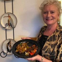 Oh What a Lucky Man he was..:) - A woman with short, curly blonde hair is holding a black plate of stir-fried vegetables, standing near a wall with decorative plates featuring artistic figures.