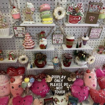 M Cafe Plus - The image shows a retail display with various holiday-themed plush pillows, ornaments, and decorative items arranged on white shelves and the floor. The display includes pink, red, and multicolored plush pillows shaped like hearts and other festive designs, along with small decorative boxes and ornaments. Signs reading 'Display Only Please Do Not Touch' are prominently placed among the items. The background features a pegboard wall with additional holiday decorations, including macarons, wreaths, and signs with Christmas motifs and messages. - Cosmetics & Beauty Supply Near Me - Arlington Heights, Illinois
