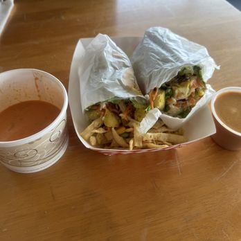 Toss’d - Love that tomato bisque with the ThaI Chicken Satay wrap - Two vegetable spring rolls wrapped in white paper are served with a side of French fries and two cups of soup, one with a reddish broth and the other with a brownish broth. The items are arranged on a light wooden table, suggesting a casual dining setting, possibly at a restaurant or takeout place. - Wraps Near Me - Temecula, California